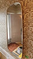 Mirror hanging on patterned wallpaper, reflecting a door and hallway area with rugs and furniture.