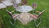 Outdoor setting showing the round glass patio table with metal frame and four matching foldable chairs with wooden slats and armrests arranged around it.