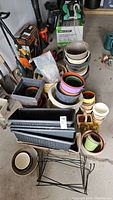 Overview of assortment of plant pots, metal stands, and rectangular planters in garage setting.
