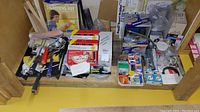 Overview of assorted plumbing and painting supplies including brushes, rollers, kits, and boxes arranged on a wooden shelf.