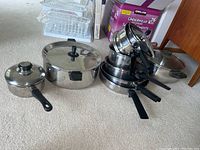 Set of eight cookware items including various sizes of pots and frying pans with black handles, mostly metal, some with lids.