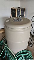 Photo of beige plastic rain barrel with wrapped spigot on top, hose coiled around base, garage background.