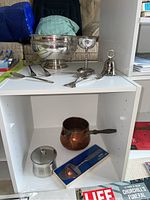 Full view of main items on white shelving including large silver plated bowl, five goblets, silver salad servers, small bell, copper pot, pewter cheese slicer in box, and stainless steel covered dish.