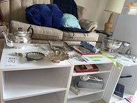 Multiple silver items displayed on white shelving including teapot, serving trays, forks, and spoons.
