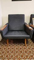 Front view of one vintage mid-century modern wood frame chair with dark fabric upholstery, wooden armrests, and legs on patterned carpet.
