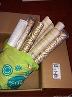Box with multiple vintage wallpaper rolls partially wrapped in plastic, showing white roll with gold speckles and beige patterned rolls.