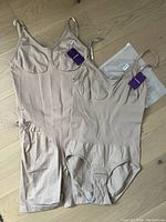 Two nude/beige Hey Shape shapewear garments with tags, one long-legged, one sleeveless, displayed on wooden floor.