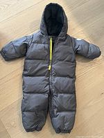 Full view of gray padded down snowsuit with hood and front zipper on wooden floor.