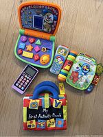 Photo showing a variety of busy baby toys including electronic learning devices, toy phones, and soft activity books arranged on a wooden floor.