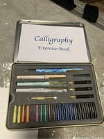 Open calligraphy set showing the exercise book, pens, nibs, and ink cartridges arranged in a foam insert inside a metal tin case.