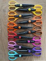 Top-down photo showing all 11 pairs of Fiskars decorative edge scissors arranged vertically on a wooden surface. Each pair has colored handles and black blade guards with unique cutting patterns and labels.