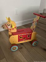 Full view of colorful wooden Janod llama ride-on toy with four rubber wheels, shape sorter on side, and red push handle at rear.