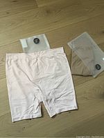 Two pairs of thigh society shorts laid out, one blush pink and one nude, with plastic packaging beside each pair.