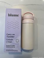 Front view of the boxed Blume carry-on insulated tumbler with the tumbler placed next to the box showing the matte white body and pastel pink handle lid.