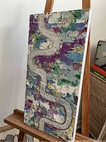 Abstract mixed media artwork on a vertical cradled wood panel shown on an easel. The panel is textured with multi-colored paint and features a wavy gold line.