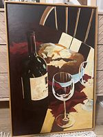 Front view of framed painting showing wine bottle, loaf of bread and two wine glasses on canvas.