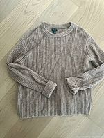 Full front view of the beige marled knit sweater laid flat showing ribbed crew neckline, long sleeves with ribbed cuffs, and hem.