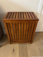 Full front view of the teak laundry hamper closed, showing vertical slats and smooth finish.