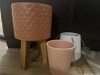 Photo shows three indoor plant pots including a tall terra cotta textured planter with wood legs, a smooth pink pot, and a white ceramic pot with drainage hole and string.