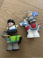 Two Fisher Price Little People knight figures each on a horse figure. One knight wears silver armor holding a blue flag on a white horse with red and blue colors. The other knight is in a white and black outfit on a gray horse with green, purple and yellow accents.