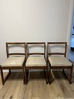Front view of three vintage solid oak dining chairs with light beige cushioned seats and rectangular backrests featuring carved detailing.