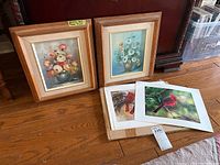 Two framed floral paintings side by side on floor, wooden frames with fabric lining and signed paintings of daisies in vases, with a box of multiple unframed bird art prints beside them.