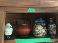 Wooden shelf displaying four Asian decorative items including an ivory egg figurine, red iron teapot, ornate round vase, and cylindrical vase with painted scene.