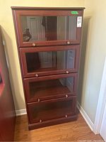 Front view of the full 4-tier wooden barrister bookcase with glass-front doors and brass knobs, placed on hardwood floor near wall corner.