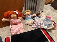 Two Cabbage Patch dolls displayed on carpet with assorted clothing beside them.