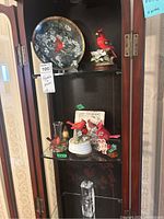 Wide view of the cabinet shelf showing various cardinal bird figurines, a decorative plate with cardinal artwork, and crystal hummingbird paperweight on the bottom shelf.