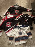Top view of all four children's hockey jerseys laid out on carpet showing the Detroit, Team Canada, CCM, and Oilers logos.