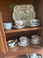 Wooden cabinet shelf with 5 floral patterned tea cups with matching saucers and a stack of small white plates, plus a square floral Myott platter behind them.
