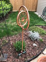 Copper garden sprinkler placed in a garden bed beside plants and mulch. Shows overall design and stake.