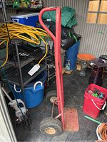 Side angled view of red metal dolly hand truck showing wheels, handle, and footplate with rust and paint chipping, located in a shed