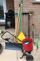 Photo showing entire lot of garden tools including seed spreader, rakes, shovels, broom, and garden hose nozzle, arranged outside on pavement against a brick wall.
