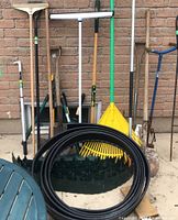 Photo showing assortment of garden tools including multiple rakes, shovels, and garden edging coils against a brick wall background.