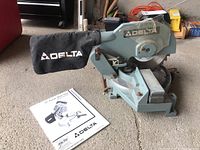 View of Delta 10-inch power miter saw with dust collection bag and instruction manual on concrete floor.