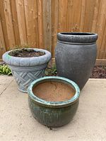 Photo of three planters outdoors against wooden fence: one gray resin planter with braided pattern, one tall dark gray resin planter with tire tread rim, and one shorter round glazed ceramic pot.