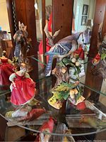 Photo showing Blue Jay bird figurine perched on branch with leaves and berries, alongside the Fair Maiden figurine in red dress reflected in the glass shelf.