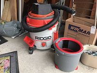 Ridgid wet/dry vacuum with orange power cord and black hose next to gray mop bucket with red wringer on garage floor.