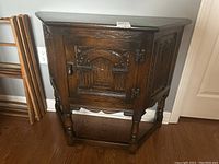 Front and side view of vintage wooden side cabinet showing carved door panel with geometric pattern, decorative metal hinges and accents, turned legs, and stretcher base.