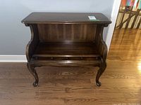 Front view of wood side table showing rectangular top, curved sides, and a drawer with four wooden knobs. Table stands on cabriole legs on wood floor.