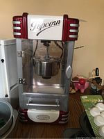 Front view of Nostalgia Electrics vintage style popcorn maker on wooden surface, showing clear glass panels, metal base with red accents, and 'Popcorn' label on top.