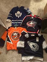 Four youth hockey jerseys laid on carpet showing Toronto Maple Leafs, NY Islanders, Columbus Blue Jackets, and Edmonton Oilers team logos and colors.