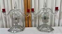 Pair of vintage clear glass double-arm candleholders shown side by side on table against curtain background. Each has a pointed finial on top and red candle holders on each arm.