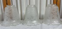 Three frosted glass lamp shades with embossed tulip floral design and ruffled edges lined up on white cloth.
