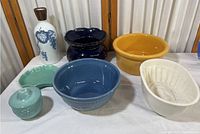 Overview photo of seven ceramic and pottery kitchen items arranged together on a table, showing bowls and dishes of various colors and shapes.