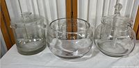 Front view of three clear glass serviceware items: two lidded jars on sides and open bowl in middle on white cloth.
