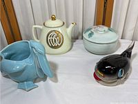 View of four pottery items on display - blue bird planter, cream teapot with gold design, small lidded ceramic container, and black bird figurine.