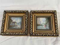 Two small square ornate framed prints shown side by side on white cloth.
